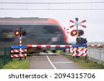Small photo of A train passes a railway crossing at high speed. Train is blurred by the speed of the train. Dutch text on the blue signs means: Wait until the red light is off, another train may come.