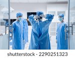 Small photo of Selective focus of a doctor's back in surgical uniform and gloves, tying a surgical cap while walking into an operating room with two medical assistants in surgical clothes standing holding door open.