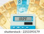The acronym IRPF referring to the Personal Income Tax in Brazil in Brazilian Portuguese written on the display of a calculator with banknotes of reais in the composition. Brazilian economy and investm