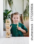 Small photo of Smiling canny cute child in green dress looking at a wooden jenga tower standing on a table