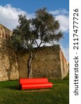 Small photo of Sanremo, Imperia, Liguria, Italy - 01 03 2022: Detail of the exterior of the Fort of Santa Tecla (1756) with a modern red bench under a tree in a sunny day