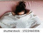 Playful young woman hiding face under blanket while lying in cozy bed, pretty curious girl feeling shy peeking from duvet, covering with white sheet, head shot close up. Top view