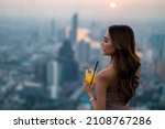 Asian woman drinking cocktail while waiting for meeting with friends at skyscraper rooftop restaurant in metropolis at summer sunset. Beautiful female enjoy outdoor lifestyle in the city at night