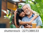 Small photo of Asian woman surprise hugging elderly father from back at outdoor garden cafe restaurant on summer vacation. Family relationship, holiday celebrating, father's day and old people health care concept