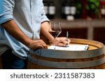 Professional man sommelier testing red wine in wine glass with tasting and smelling at wine cellar with wooden barrel in wine factory. Winery liquor manufacturing industry and winemaker concept.