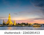 Small photo of At dusk,the silhouette of a Thai Long Tail boat drifting past the golden pagodas of the temple complex,as they point towards clear sunset skies,seen from opposite side of Bangkoks main river.