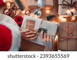 Top view hands of woman preparing handmade Christmas gift, Female wearing Santa hat making beautiful present gift box on the table, Christmas eve preparation