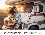 Hipster beautiful woman playing guitar with big smile on her face. Nomadic and camper lifestyle concept. Warm sunset light. 