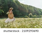Small photo of A young woman in a white sundress, a wreath of daisies with a large wicker bag on her shoulder is walking through a field of daisies, against the background of the forest, the sunset light