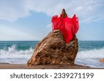 Small photo of Red dress sea woman. Yappy woman with flowing hair in a long flowing red dress stands on a rock near the sea. Travel concept, photo session at sea