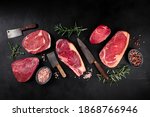 Meat flat lay. Various cuts of meat, overhead flat lay shot with knives, salt, pepper, and herbs, on a black slate background