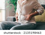 Small photo of Midsection of woman hands holding her belly fat sitting on sofa at home. Caucasian lady grabbing excessive fat on her abdomen. Young female pinching on her fatty obese waist