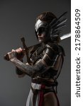 Small photo of Attractive valkyrie from past with make up holding sword on her shoulder against grey background.