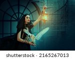 Small photo of Gothic fantasy woman witch in black dress cape, hood. Girl princess dark magician holds vintage lamp lantern in hand. Lady elf queen with white barn owl bird. Art photo night royal room. Fabric flies
