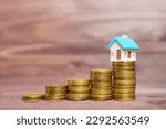 Small photo of Colorful toy house figure standing on top of a pile of coins, all set against a wooden background. Personal finance, investing, real estate and wealth management related concept.