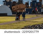 Small photo of Two adult women with young baby and stroller walking at "Boxcar Park" in Everett, Washington on a sunny brisk winter afternoon sunset. A industrial background and shipyard behind