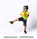 Small photo of Kid in sunglasses, green hat, yellow t-shirt, denim shorts, khaki sneakers. Jumping, drinking cocktail from plastic bottle like pineapple through tubule. Isolated on white. Full length, copy space