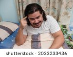 Small photo of A lazy and smart aleck bearded guy in his 30s thinks he is clever pointing to his brain while sitting on the bed after a nap.