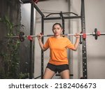 Small photo of A pensive asian woman performing a set of barbell squats from a home workstation. Possibly being a bit nonchalant or uninspired to work out.