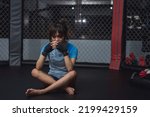 Small photo of A sad female Muay Thai novice squats down inside the boxing ring cage as she thinks about the game wherein she lost.