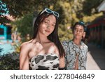 Small photo of A confident young woman with a enamored and infatuated man admiring her from behind. Outdoor resort scene.