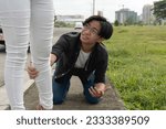 Small photo of A pitiful young man kneels down and grabs his girlfriend's leg in a desperate attempt to prevent her from leaving him. Breaking up with a weak or insufferable guy