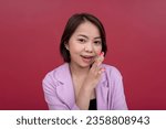 Small photo of A chatty young asian woman in her mid 20s spreading hearsay or gossip secretly. Wearing a pink blazer and black tube top. Studio shot with burgundy background.