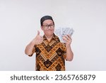 Small photo of A middle aged man making a thumbs up sign while holding a lot of cash in his hand, flaunting his wealth or earnings. Wearing a batik shirt and songkok skull cap. Isolated on a white background.