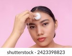 Small photo of A pensive young Asian woman using a cotton ball to apply toner or astringent on her forehead. Isolated on a pink background.