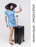 Small photo of A full-length portrait of an Asian woman tourist capturing a selfie with puckered lips while standing with her suitcase.