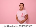 Small photo of A smiling mixed race woman in pink T-shirt with pink satin ribbon symbolizing International Breast Cancer Day, expressing solidarity and support for breast cancer patients and survivors. 1 st october