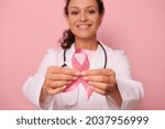 Small photo of Soft focus on Breast Cancer Awareness Pink Ribbon, in hands of blurred mixed race woman doctor in white medical coat, isolated on colored background. 1 st October, World Day of fight breast cancer