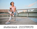 Small photo of Happy delighted Caucasian female athlete sportswoman in pink t-shirt with smartphone holder and terry wristbands rests after morning jogging at dawn sitting on a wooden bench in an urban environment