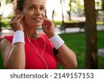 Small photo of Attractive middle aged active woman, sportswoman putting on earphones, wearing bright red t-shirt and white terry wristbands, getting ready for outdoor workout and morning run