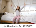 Small photo of Caucasian beautiful little girl waking up in the morning, stretching her arms upwards, smiling cutely, looking at camera, sitting on the comfortable double bed with white bedsheets in her bedchamber