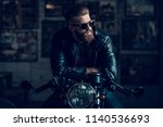 Young Bearded Biker Sitting on Motorcycle in Garage. Indoor Garage. Young Mechanic in Garage. Parts of Motorcycle. Man in Checkered Shirt. Man on Vintage Bike. Biker Lifestyle Concept.