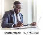 Handsome Afro American businessman in classic suit and eyeglasses is using a laptop and making notes while working in office