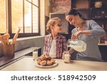 Cute little girl and her beautiful mother are smiling while drinking milk and eating muffins in kitchen. Mother is pouring milk