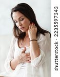 Small photo of Middle aged woman sitting on a white bed in a bedroom at home touching chest with her hands while having a heart pain and feeling unwell. Suffering from stress, hypertension or cardiac arrhythmia