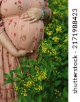 Small photo of A young woman in pink printed tight-fitting dress embraces her belly with a future baby with trepidation and tenderness. yellow flowers . selective focus" " follow focus" " shallow depth of field"