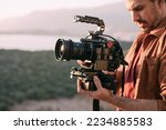 A young Caucasian man with a professional camera in his hands on a mountain by the sea.  The operator holds a video camera, shoots a sunset landscape, mountains, sea, setting sun