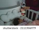 Small photo of unknown woman mother adjusting and setting up Surveillance security camera on baby bed at home in bedroom watching small child in cradle while sleeping in dark room at night