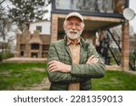 One modern senior caucasian man portrait of male grandfather with beard and hat cap stand in front of his house in day wear jacket happy confident copy space arms crossed