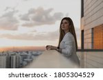 Young pretty woman dressed on white shirt looking at city from balcony, enjoying the sunset at terrace. Shot of beautiful young woman standing on the balcony and looking at the view. Relax, dreaming