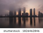Modern high rise condo and business buildings at waterfront covered with low clouds, fog, haze, smoke at sunrise sunset hour reflecting in still water surface. Hot real estate market concept.