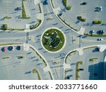 Direct overhead aerial view of a 4-way roundabout in a city center, all sides surrounded by lightly filled parking lots.