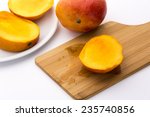 Small photo of Golden yellow third of a trisected mango. Its ripe fruit flesh bedewing the wooden cutting board beneath. The remaining two thirds on a white plate next to an entire mango. White background.