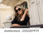 Beautiful smiling blond woman in elegant luxury sunglasses posing on the street. European background. Perfect wavy hairs.