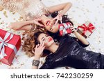 Two smiling gorgeous  women  in trendy sequin dress lying on white floor with shining golden confetti and red gift boxes . Celebrating new year or birthday party. Showing peace.