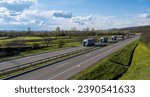 Small photo of Caravan or convoy of trucks in line on a country highway. A convoy of trucks (lorries) heading down the highway between meadows and dense forests. White clouds are covering the sky.